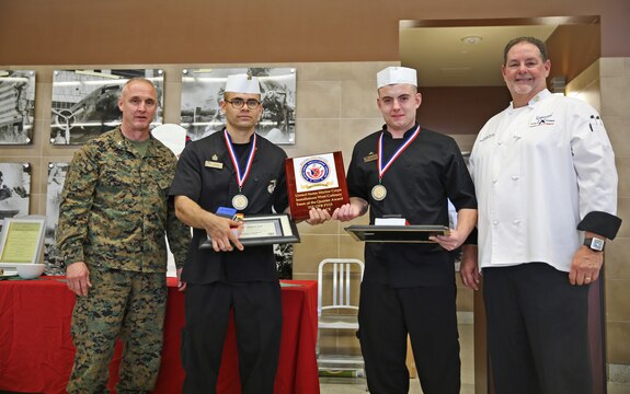 Sgt. Hugo P. Zepeda and Lance Cpl. Justin Gordon, food service specialists with Headquarters Squadron, Marine Corps Air Station Yuma, pose for a photograph with Col. Jeff Arruda, left, chief-of-staff, Marine Corps Base Camp Pendleton, Marine Corps Installations-West and Donovan Brown, right, head chef with Sodexo, after taking first place honors at the MCI-West Chef of the Quarter culinary competition here, Dec. 3. 