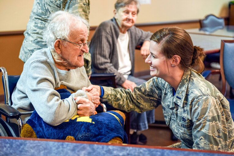 An airman kneels and smiles while talking to a World War II vet sitting in a wheelchair.