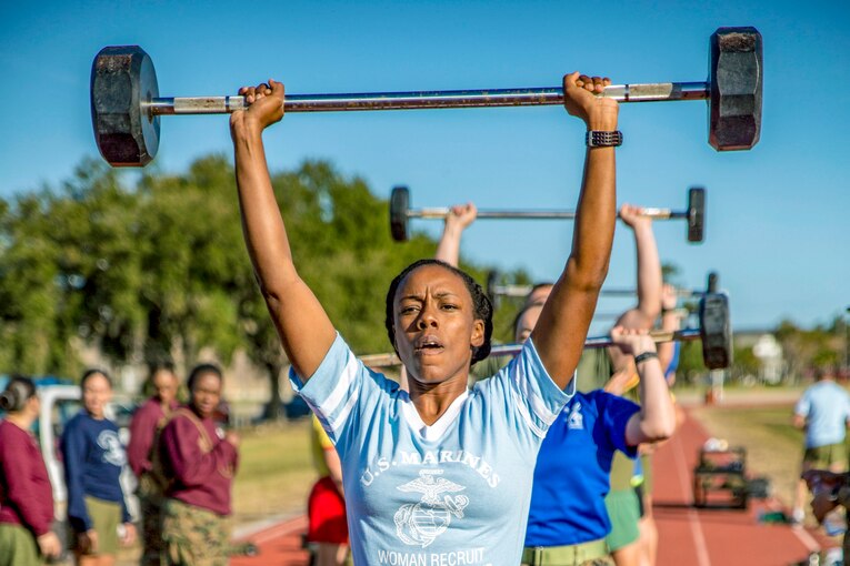 Marines stand in line outside and  lift  a barbells over their heads.
