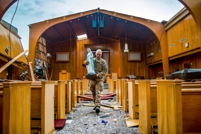 A chaplain carries a statuette of a woman down a walkway of a damaged church, flanked by pews.