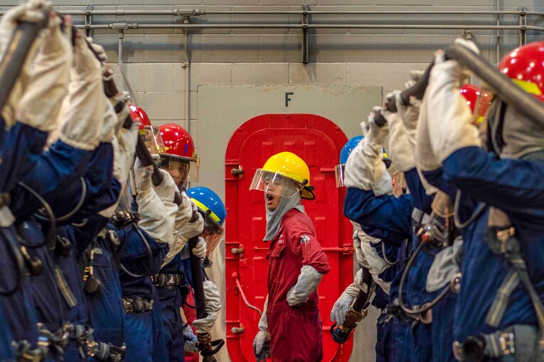 An instructor in red shouts orders to two lines of recruits in blue uniforms and red hard hats holding up hoses