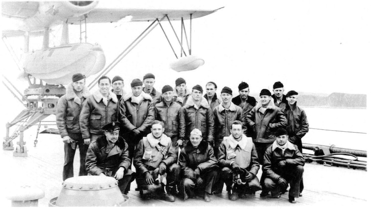 Aviation Radioman 2nd Class Harrison D. Miller (back-center) aboard the flight deck of the USS New Mexico (BB-40) with fellow flight crews for OS2N-1 ?Kingfisher? observation/scout float planes.  Miller later received the Distinguished Flying Cross for his heroic actions on 31 January 1944 when U.S. forces invaded Kwajalein Atoll. At 1522 local time, his plane was hit by enemy anti-aircraft fire from Ebeye Island, an enemy float plane base at the time. The Kingfisher suffered serious damage and was forced to make an emergency water landing in the lagoon due to high octane aviation fuel leaking into the bilges in the cockpit, filling it with fuel fumes presenting a critical fire hazard.  The pilot, Navy Lieutenant Forney O. Fuqua was mortally wounded by the enemy fire and instructed the Miller, in the rear cockpit of the Kingfisher, to bail out.  Miller elected to stay with the plane and to attempt a water landing himself from the rear cockpit.  With no prior flying experience, no flight instruments and only an emergency control stick in the rear cockpit, he successfully made a water landing from the back seat of the Kingfisher.  After landing, Miller got out of the rear cockpit onto the wing and made his way to the front cockpit and turned off the Kingfisher?s engine and started to unbuckle the pilot?s parachute and harness; but before he could get him unbuckled, the plane capsized due to the loss of the outboard pontoons during the landing.  Miller made numerous attempts to rescue the pilot and remove him from the cockpit, diving under the gasoline covered waters but was unsuccessful.