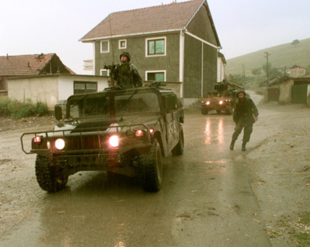 Soldiers of Alpha Company, 1st Battalion, 77th Armored Regiment wait in the rain by their Humvees as their fellow soldiers search for automatic weapons in the town of Zitinje, Kosovo, on July 26, 1999. The soldiers of the 77th are deployed to Kosovo as part of KFOR. KFOR is the NATO-led, international military force in Kosovo on the peacekeeping mission known as Operation Joint Guardian. 