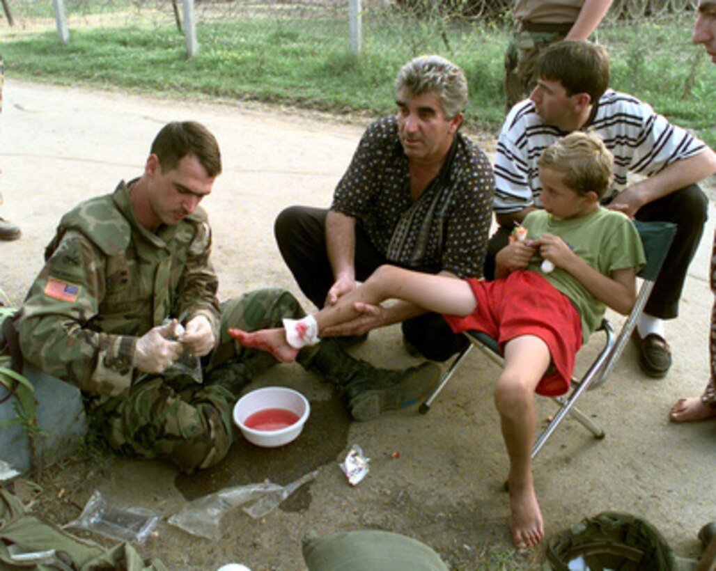 Sgt. Ray Lester, U.S. Army, sits on the ground as he works to sterilize and bandage a dog bite on the leg of Gezim Ademi in the town of Vitina, Kosovo, on July 20, 1999. Lester is attached to the 1st Battalion 77th Armor Regiment which is deployed to Kosovo as part of KFOR. KFOR is the NATO-led, international military force in Kosovo on the peacekeeping mission known as Operation Joint Guardian. 