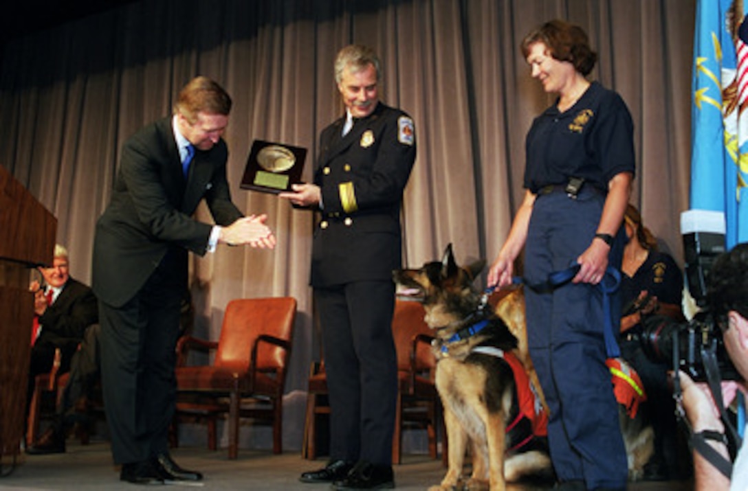 Secretary of Defense William S. Cohen (left) applauds the search dog Nike and his handler Susan Mingle (right) after presenting Assistant Chief Mark Wheatley (center) of the Fairfax County Urban Search and Rescue Team a plaque in ceremonies at the Pentagon on June 27, 2000. Cohen lauded the team for their continuing tireless work and selfless dedication in providing humanitarian assistance to victims of disasters worldwide. Wheatley accepted the honor on behalf of the approximately 100 team members and three search dogs in the audience. 
