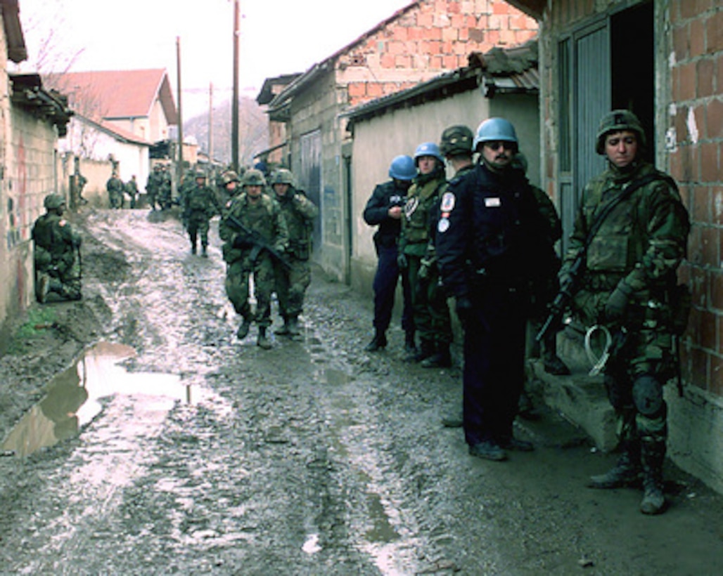 Soldiers from the U.S. Army's Bravo Company, 3rd Battalion, 504th Parachute Infantry Regiment, and United Nations' police move down a muddy alley way in Mitrovica, Kosovo, as they conduct a house-to-house search for weapons on Feb. 21, 2000. The soldiers are attached to the 82nd Airborne Division, Fort Bragg, N.C., and are deployed to Kosovo as part of KFOR. KFOR is the NATO-led, international military force in Kosovo on the peacekeeping mission known as Operation Joint Guardian. 