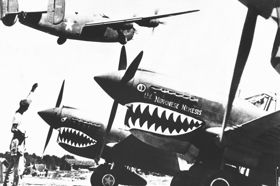 1940's -- Landing wheels recede as this U.S. Army Air Forces Liberator bomber crosses the shark-nosed bows of U.S. P-40 fighter planes at an advanced U.S. base in China.  An American soldier waves good luck to the crew, off to bomb Japan.  1943  Air Force Photo