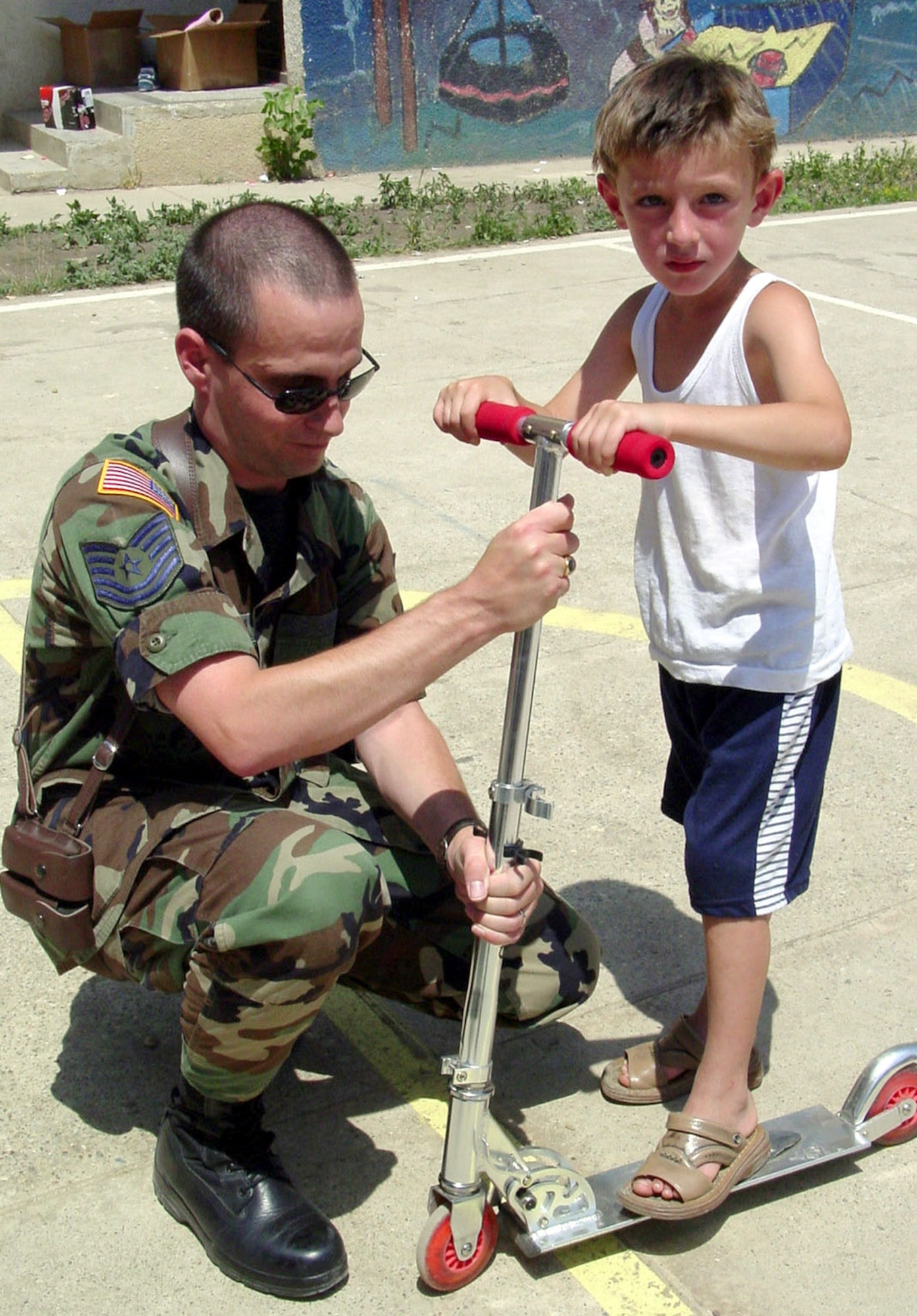 PRISTINA, Kosovo -- Tech. Sgt. Ernie Kindle readies a new scooter for 5-year-old Aragon's first scooter ride at the Pristina Mother and Child Center here.  Sergeant Kindle is the Kosovo Force disbursing noncommissioned officer and is deployed from Wright-Patterson Air Force Base, Ohio.  (U.S. Air Force photo by Master Sgt. Tim Helton)