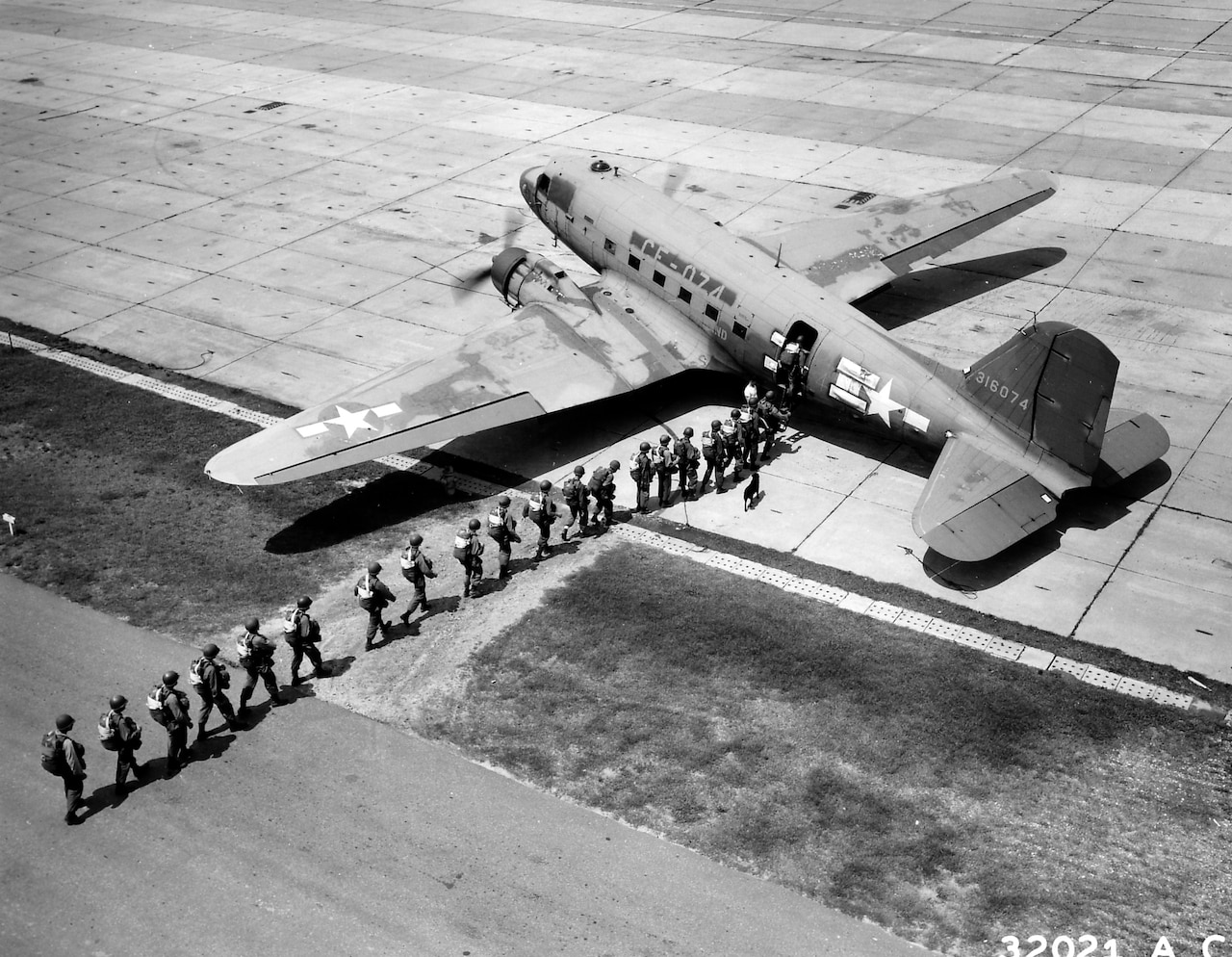 In a black and white photo a line of paratroopers board an aircraft on a runway.