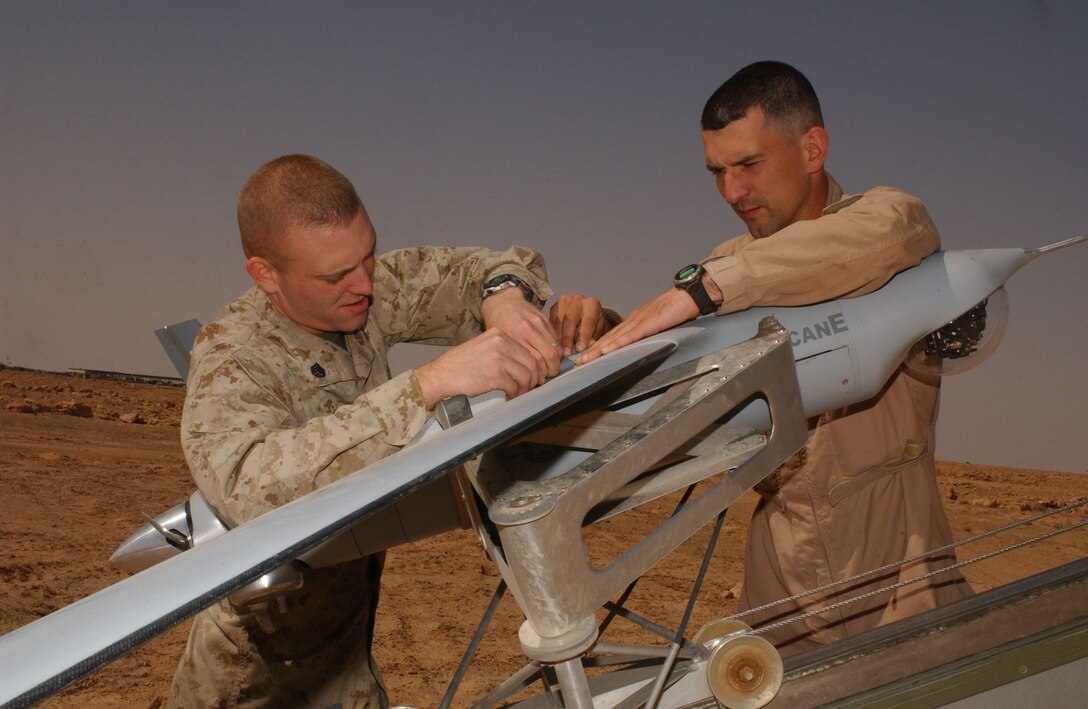 AL ASAD, Iraq--Staff Sgt. Robert D. Custer, intelligence analyst, and Cumberland, Md., native, and Martin Susser, a native of White Salmon, Wash., who works for Insitu Group, and a ScanEagle operator, prepare the unmanned aerial vehicle for flight.  They are part of a small detachment of Marines and civilians making up Marine Unmanned Aerial Vehicle Squadron 2, 2nd Marine Aircraft Wing, from Cherry Point, N.C.  The detachment is deployed here to provide intelligence, surveillance, and reconnaissance in western Iraq.  ScanEagle weighs approximately 40 pounds and has a 10-foot wing span.  The aircraft operates with a small engine, requiring a small amount fuel.  Its 4-foot frame can remain airborne for more than 10 hours.  Each drone is launched using a catapult system, which makes it runway independent and perfect for forward operating forces.  Using the Global Positioning System, it is retrieved with a skyhook where the UAV catches a small, suspended rope. Saving lives is the number one mission for this small detachment.  As they collect information on potential targets, the leaders on the ground decide what effective measures to take to minimize the risks against friendly forces.  Spending almost 15 hours a day gathering information, the analysts provide a solid foundation to make tactical decisions.