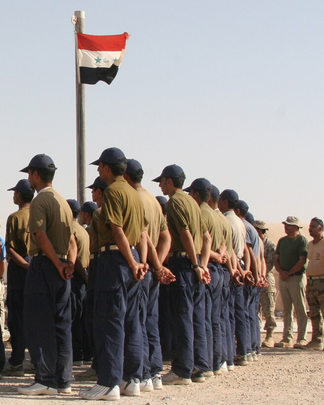 AL ASAD, Iraq - Iraqi men stand beneath their national flag during their graduation ceremony Aug. 24 as they listen to the introduction being given by the academy director, Chief Warrant Officer Scott Reinhardt.  More than 100 Iraqi police officers graduated from the advanced course, it was the last class to attend the academy here.