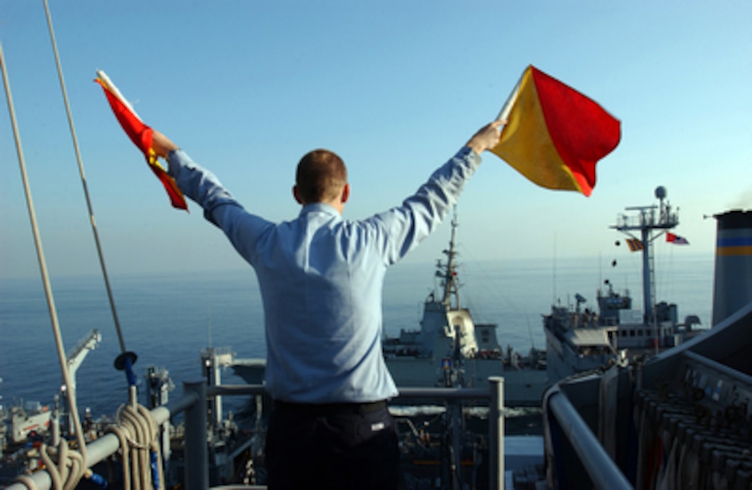 Navy Seaman Ryan Ruona uses semaphore flags to signal another ship alongside the aircraft carrier USS Theodore Roosevelt (CVN 71) during replenishment at sea on Nov. 29, 2005. Roosevelt and its embarked Carrier Air Wing 8 are conducting operations in the Persian Gulf. 