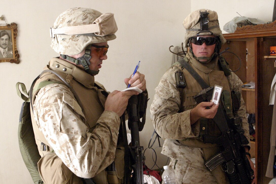 FALLUJAH, Iraq - Lance Cpl. Dan Williams, an intelligence analyst with 1st Battalion, 6th Marine Regiment, jots down identification information on a local resident, as infantryman Cpl. Eric Beaverson holds up an ID card.  Williams, a 24-year-old Murrysville, Penn. native, is currently deployed to Iraq's Al Anbar province, where he assists his Camp Lejeune, N.C.-based unit acquire information on insurgent whereabouts.