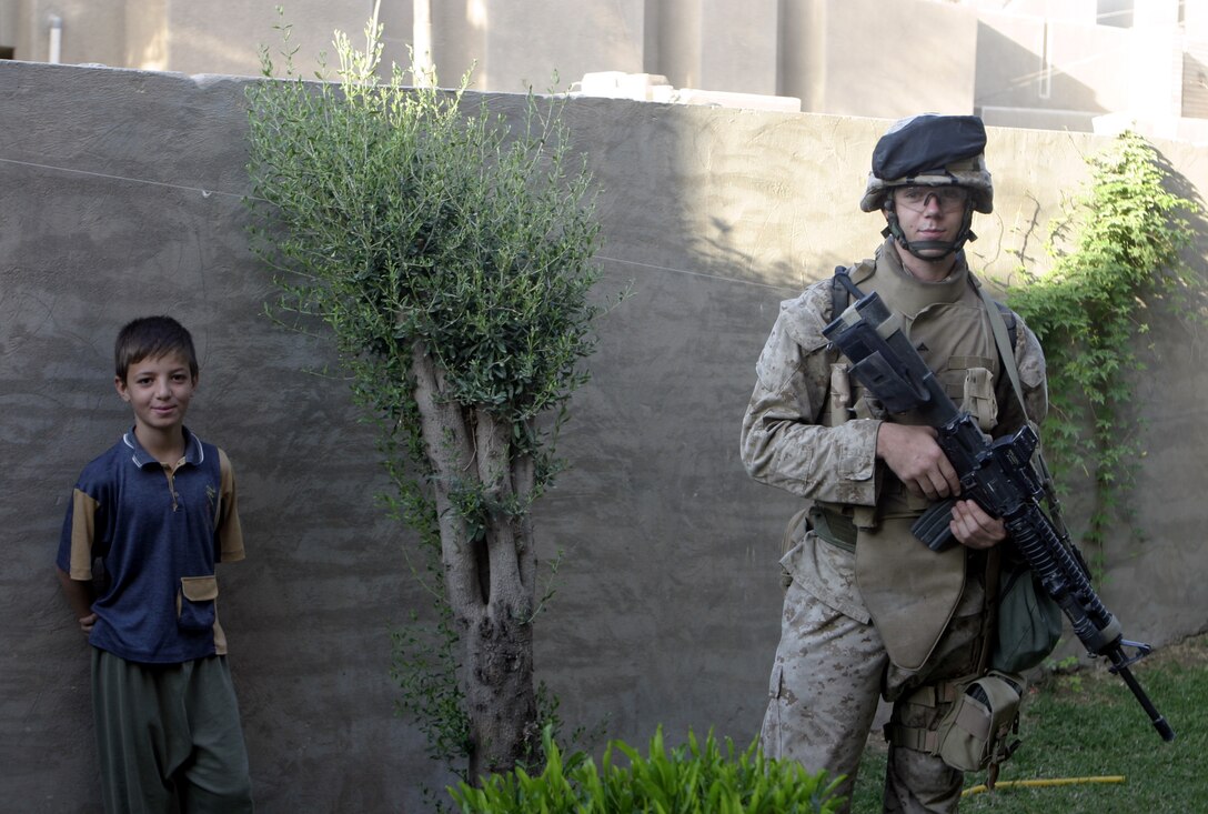 FALLUJAH, Iraq - Lance Cpl. Raleigh Krigbaum, an infantryman with 2nd Platoon, Company B, 1st Battalion, 6th Marine Regiment, provides security outside a home here July 7 while a child looks on.  Company B personnel conducted Operation Hard Knock alongside Iraqi Security Forces here, during which the troops wired off entire neighborhoods and searched through each house for concealed weapons and explosives.