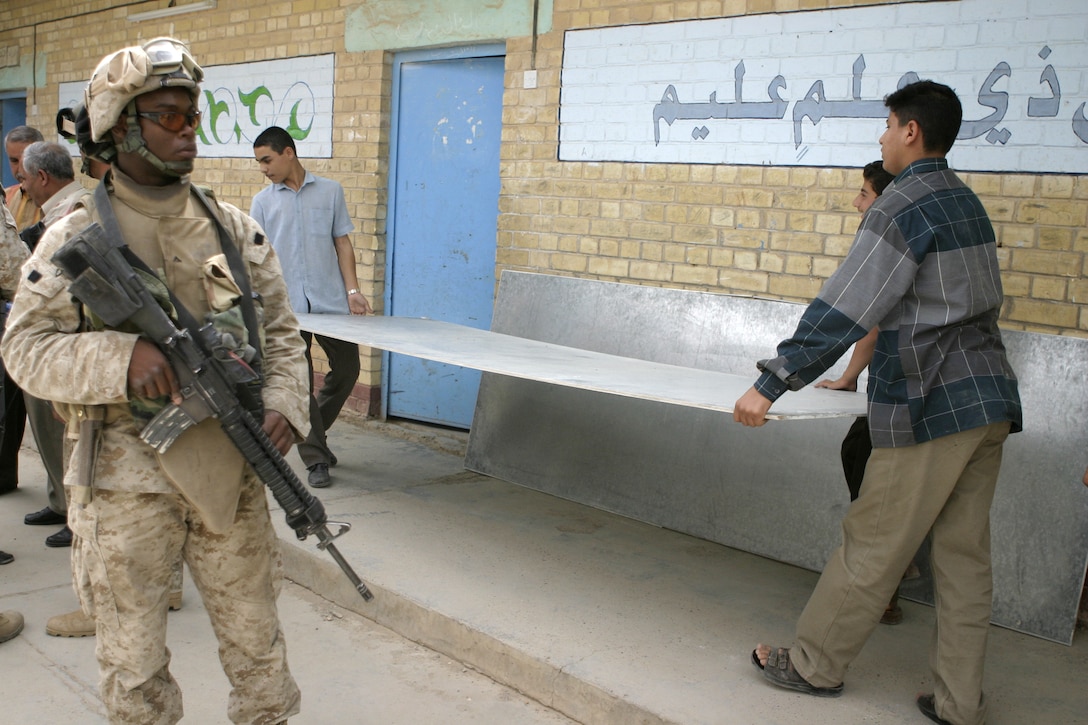 FALLUJAH, Iraq - Lance Cpl. Brandon C. Dinkins, a machine gunner with 1st Platoon, Company C, 1st Battalion, 6th Marine Regiment, provides security at a school here May 12 as Iraqi students in the background help carry out an old blackboard from their classroom, while Iraqi soldiers hang up a new one.  The 19-year-old Shreveport, La. native, his unit, and Iraqi Security Forces are currently conducting 'Operation Blackboard,' a joint civil-military mission during which Iraqi soldiers re-supply Fallujah's schools and continue winning the citizens' trust.