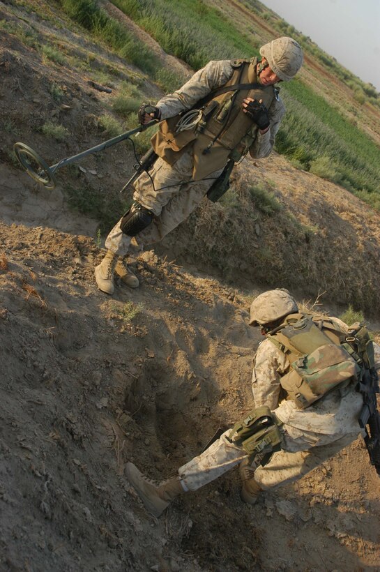 AR RAMADI, Iraq (Aug. 15, 2005) - Lance Cpl. Brandon C. Jordan, a combat engineer with 4th Platoon, Company A, 1st Combat Engineer Battalion, which is in direct support of 1st Battalion, 5th Marine Regiment, stands by with his metal detector in hand while his partner, Lance Cpl. Matthew F. Tripp, digs for weapons buried in a field on the outskirts of the city here during an early-morning mission Aug. 15. Jordan, Tripp and other combat engineers uncovered several large weapons caches while supporting Operation Bullard II. First Battalion, Fifth Marines carried out the operation, which started before daylight and lasted until midday, in and around a small community on the edge of volatile Ramadi in an effort to capture insurgents and weapons. Combat engineer Marines discovered a mortar tube, more than 200 artillery shells in sizes ranging from 60 mm to 155 mm, four .50 caliber machine gun barrels plus more than 2000 rounds of ammunition and approximately 1000 7.62 mm rounds, 600 of which were armor piercing. Infantrymen with the battalion searched houses, vehicles and personnel and captured 14 insurgents. Photo by: Cpl. Tom Sloan