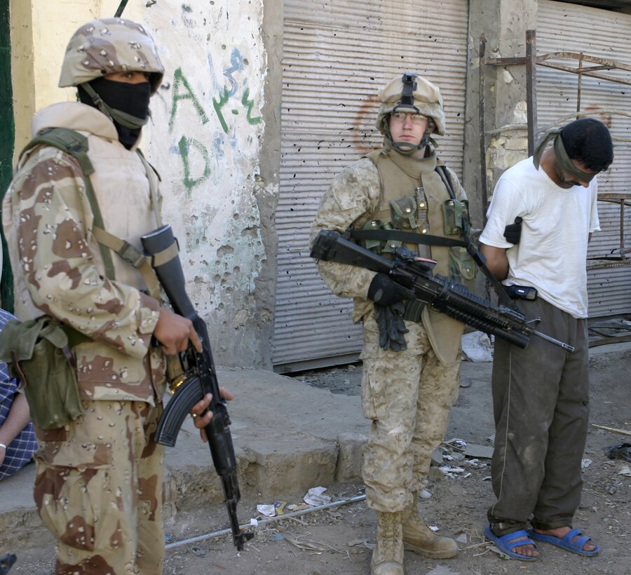 FALLUJAH, Iraq - An Iraqi soldier guards a detainee alongside Lance Cpl. Matt R. Walters, an assistant team leader with 3rd Squad, 3rd Platoon, Company C, 1st Battalion, 6th Marine Regiment, at the Farwat bakery here.  Iraqi Security Forces and Company C personnel conducted a raid on the bakery, detained nine suspected insurgent supporters, and confiscated a pistol and numerous forms of anti-Coalition forces propaganda.