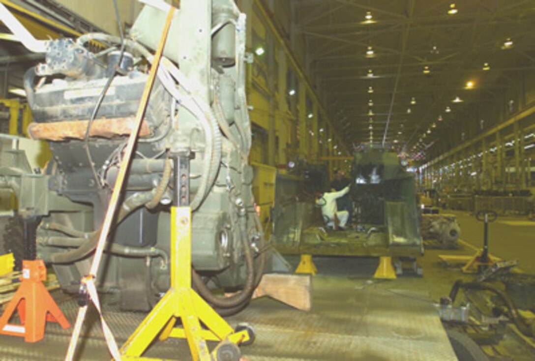 The engine of the M9 Armored Combat Earthmover sits on floor jacks outside of the vehicle. Behind the engine, Art Torres, heavy mobile equipment mechanic, works on the vehicle's hydraulic system.