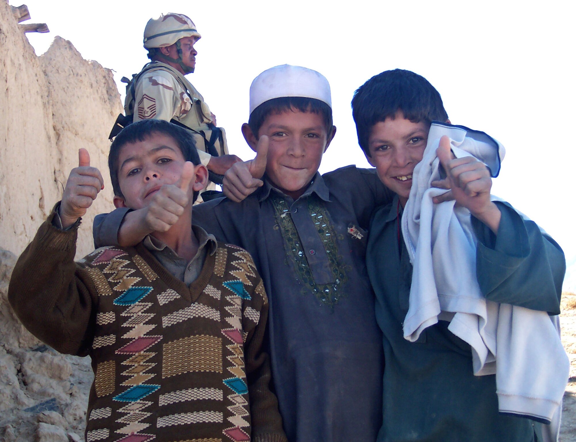 PARWAN PROVINCE, Afghanistan (AFPN) -- Local Afghan boys give a "thumbs-up" as Senior Master Sgt. John Jones provides protection in the background during an Adopt-A-Village visit. Airmen from nearby Bagram Airfield distributed more than 1,000 pounds of supplies to the local village. Sergeant Jones, a native of Winnfield, La., is deployed from Elmendorf Air Force Base, Alaska. (U.S. Air Force photo by Capt. Robert Lunden) 