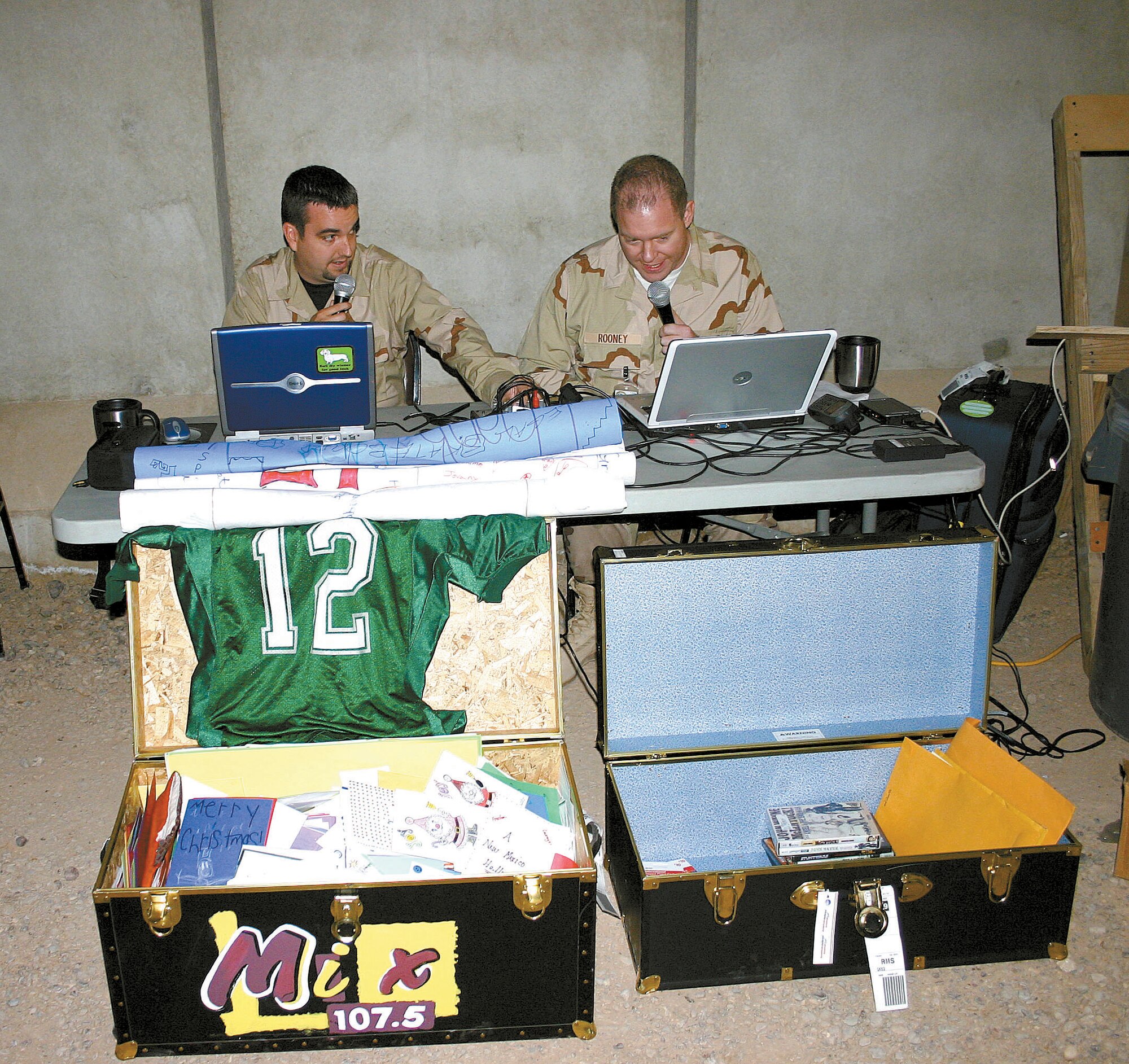 Clovis radio personalities Duffy Moon (left) and Steve Rooney (right) perform their usual morning show live from Balad Air Base, Iraq, outside the recreation center. Deployed Airmen from the 27th Fighter Wing were given the opportunity to send live holiday greetings back home. In front of them are two footlockers they brought with them containing messages, CDs, DVDs and phone cards donated by members of the local communities. (photo by Staff Sgt. Craig Seals)