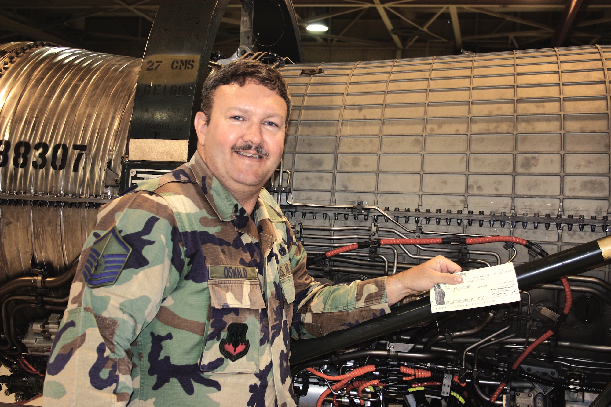 Master Sgt. Ken Oswald, 27th Fighter Wing Safety, Cannon Air Force Base, N.M., displays a check as he stands in from on an F-16 compressor case. Sergeant Oswald received $7,000 on May 4 for improving the procedure for removing the compressor's blade lock, a procedure that prevents future damage and result in saving the Air Force $50,342. He will represent Air Combat Command for the 2006 Air Force Exceptional Innovator Award. (Air Force photo by Greg Allen)