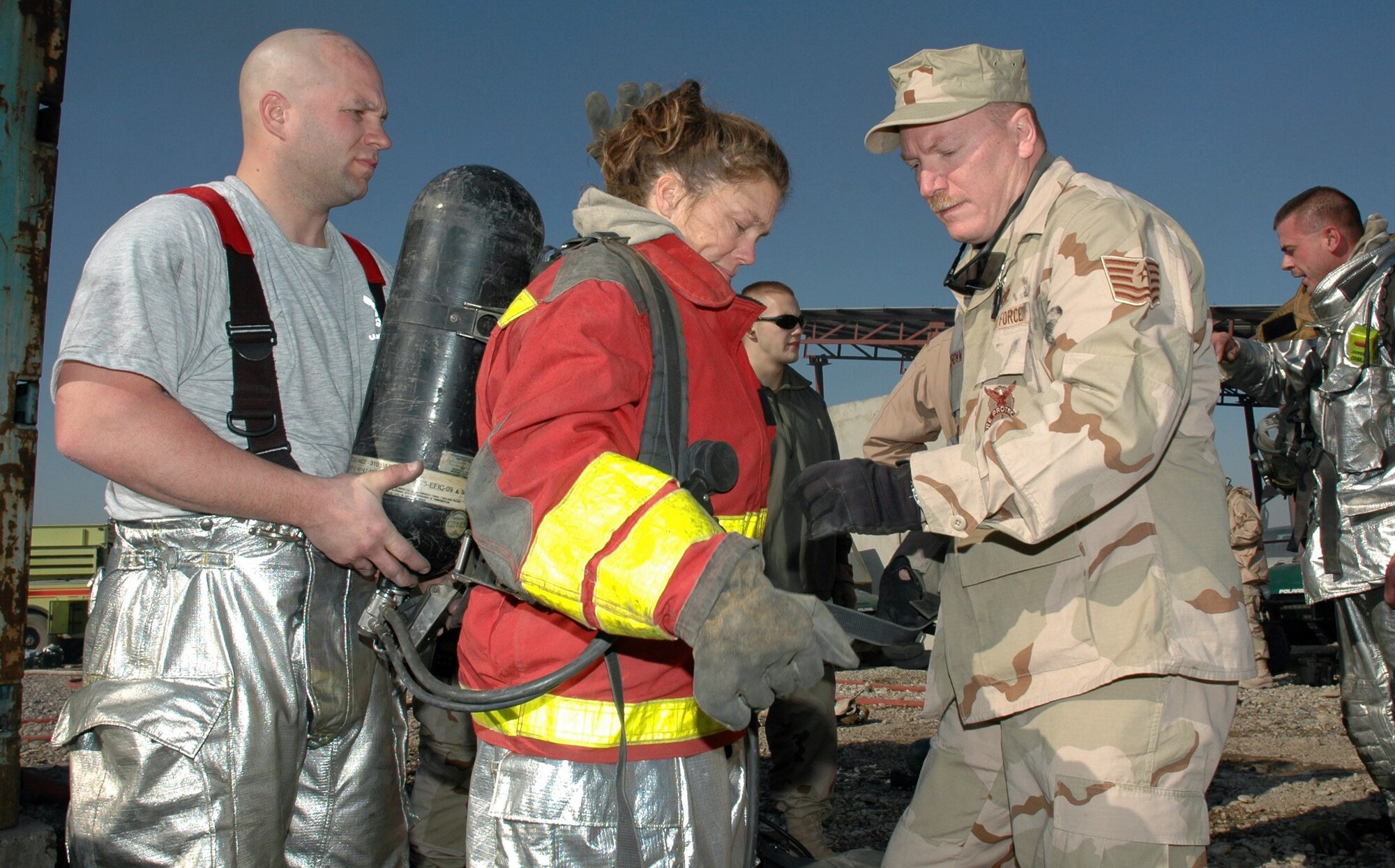 KIRKUK AIR BASE, Iraq (AFPN) -- (From left) Tech. Sgt. Matt Lyons holds a compressed air tank for Capt. Cindy Lang as Tech. Sgt. Charles Brown assists her with the donning of the protective fire suit. Sergeant Lyons is the assistant chief of training, and Sergeant Brown is a battalion fire chief here. Captain Lang is a critical care nurse who volunteered to experience the smoke simulation burn trainer. (U.S. Air Force photo by Tech. Sgt. Ken Sloat) 