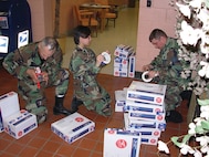 From left: Tech. Sgt. Thomas Vera, Staff Sgt. Millicent Costiniano, and Tech. Sgt. Jason Rootes,  from the 5th Communications Squadron, package supplies of pens, pencils, notebooks and more to send to Master Sgt. Michelle Rootes, 5th Medical Operations Squadron and Jason’s wife, who is deployed to Afghanistan and volunteering to help a local school. (courtesy photo)
