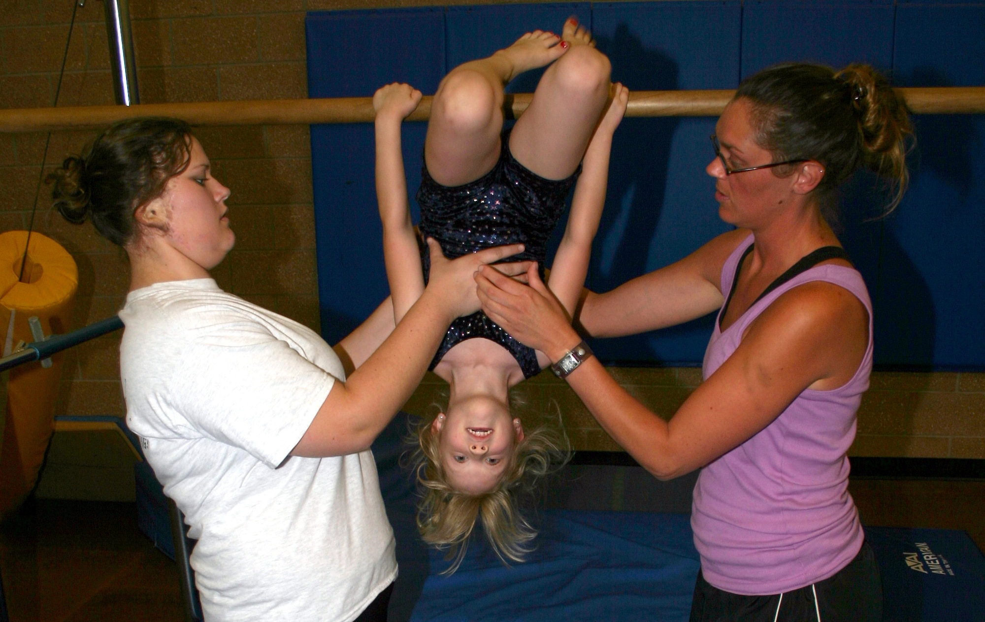 KinderGym teachers Deann Dangelmayr (left) and Sarah Bristow assist student Meredith Johnson in completing the gymnastics movement “skin the cat” on the 
uneven bars at the Madrigal Youth Center Tuesday. The center offers various classes in different skill and age levels, for information call 676-2342. (U.S. Air Force photo/Senior Airman Jacque Lickteig).