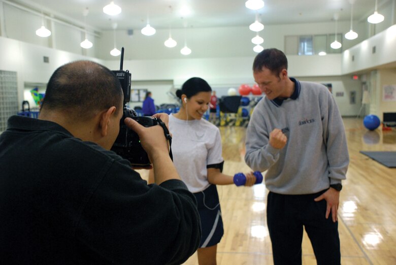 MCCHORD AIR FORCE BASE, Wash. -- Base photographer Abner Guzman, 62nd Communications Squadron, takes pictures of Staff Sgt. Irene Noriega, 62nd Airlift Wing, performing a set of dumbbell curls under the direction of personal trainer Staff Sgt. Ronald West, 62nd Services Squadron, for a Northwest Airlifter feature story recently at the fitness center annex. (U.S. Air Force Photo by  Staff Sgt. Zachary Knittle )