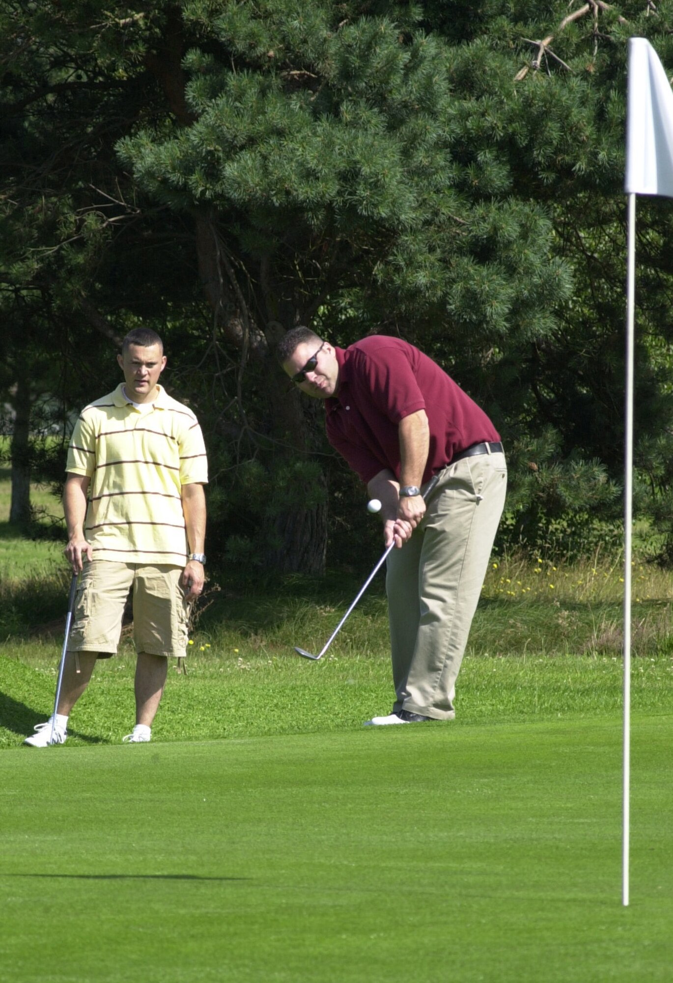 Chad Cox, playing for the Defense Courier Service, chips onto the 17th green in the intramural golf league at Breckland Pines Golf Club July 31, while teammate Shaye Melton looks on. (Air Force photo by Gary Rogers)                      