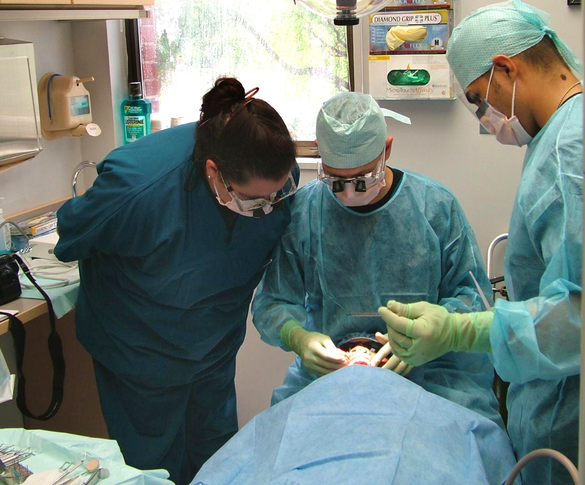 Capt. (Dr.) Bradford Allen, center, performs periodontal surgery on a patient while discussing surgical options with Maj. (Dr.) Eddi Graham (left), the program’s periodontist. The doctors are assisted by Airman 1st Class Harold Gomez (right), dental assistant. (U.S. Air Force photo by Maj. [Dr.] Dennis Holt)
