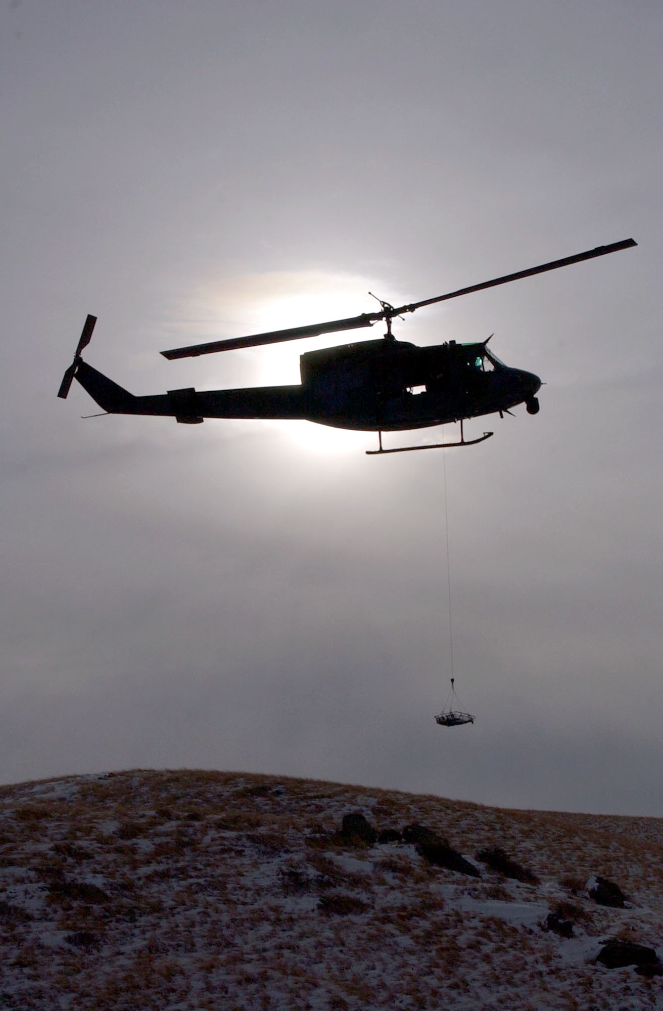 The silhouette of a UH-1N Huey helicopter from the 40th Helicopter Squadron hovers over the Montana skyline near the Rocky Mountain Front during a search and rescue training sortie Nov. 19. The stokes litter, a stretcher designed to transport people from the ground to the helicopter while in the air, is slowly lowered by the on-board flight engineer for a simulated search and recovery mission. (U.S. Air Force photo/Senior Airman Eydie Sakura).            