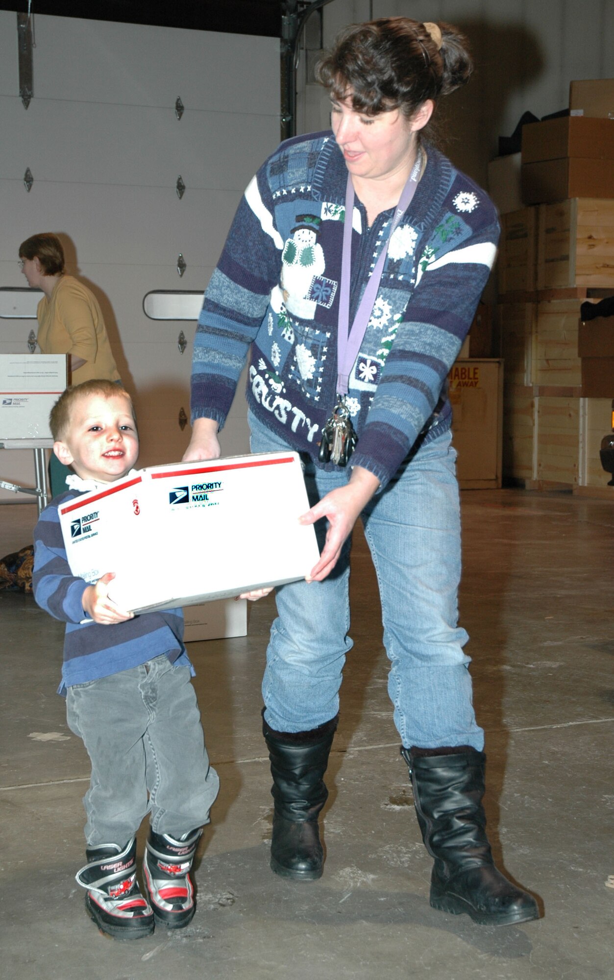 Little Warrior Danny, 3, with assistance from Connie Wright, transport a care package across the traffic management office Dec. 1. Roughly 400 care packages were mailed the same day to deployed servicemembers from Montana. (U.S. Air Force photo/Airman 1st Class Dillon White)