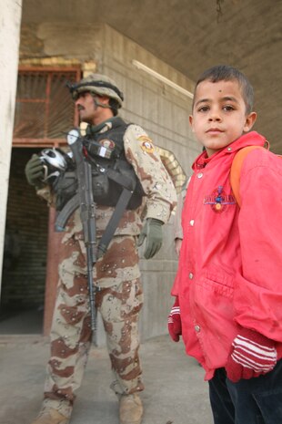 A young Iraqi child poses for a picture while an Iraqi army soldier with 1st Battalion, 3rd Brigade, 1st Iraqi Army Division stands watch at a school near Abu Hazim Dec. 8. MTT 1-3-1 is currently deployed within Iraq?s Al Anbar Province to help develop Iraqi Security Forces, facilitate the development of official rule of law through democratic government reforms and continue the development of a market based economy centered on Iraqi reconstruction.