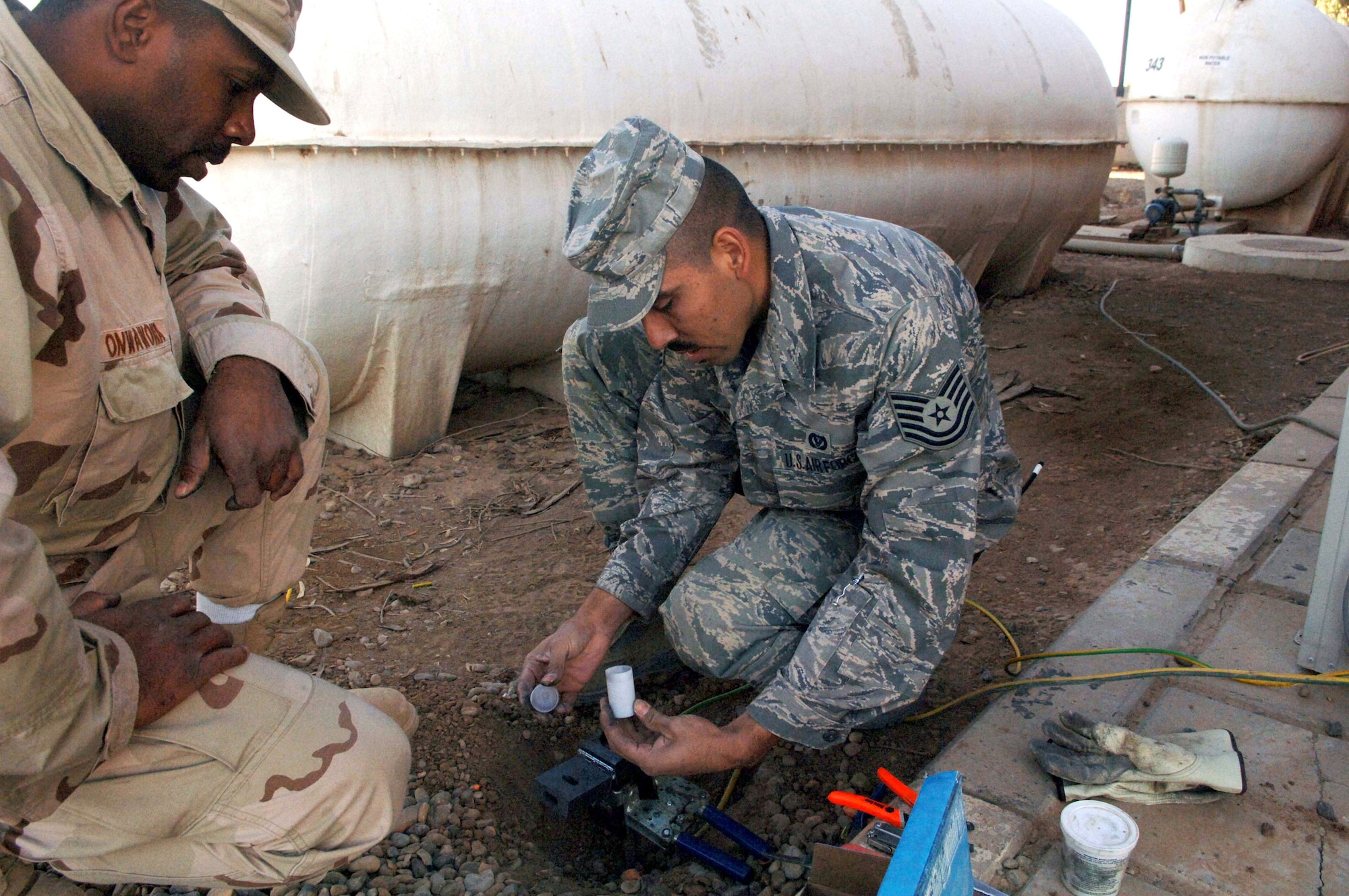 Staff Sgt. Eduardo Onwawoma watches Dec. 11 as Tech. Sgt. Victor Magana pours welding powder into an exothermic welder while working at Balad Air Base, Iraq. The welder will be used to fuse two pieces of ground wire to a grounding rod, preventing static electricity from damaging nearby communitcaion lines. Sergeants Onwawoma and Magana are electricians with the 332nd Expeditionary Civil Engineer Squadron. Sergeant Magana is deployed from Barksdale Air Force Base, La., and Sergeant Onwawoma is deployed from Yokota Air Base, Japan. (U.S. Air Force photo/Senior Airman Terri Barriere)