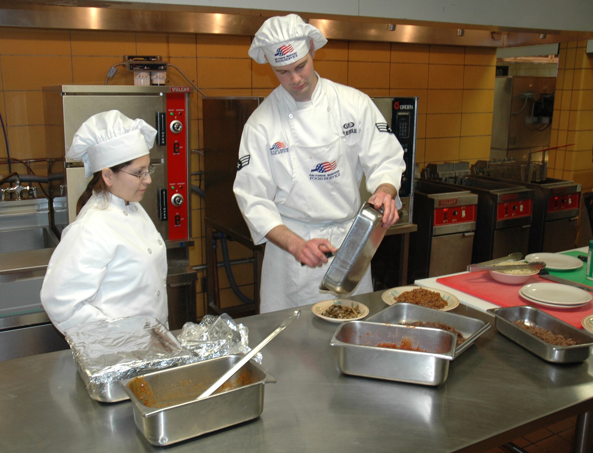 MINOT AIR FORCE BASE, N.D. -- (From left) Sous-chef Jenna Seaboy looks on as Senior Airman Travis Keele puts the finishing touches on his entree. Airman Keele and his sous-chef went with a classic Italian approach combined with some old fashioned American cuisine: Spaghetti with sautéed mushrooms and sloppy joes. (U.S. Air Force photo by Airman 1st Class Wesley Wright)