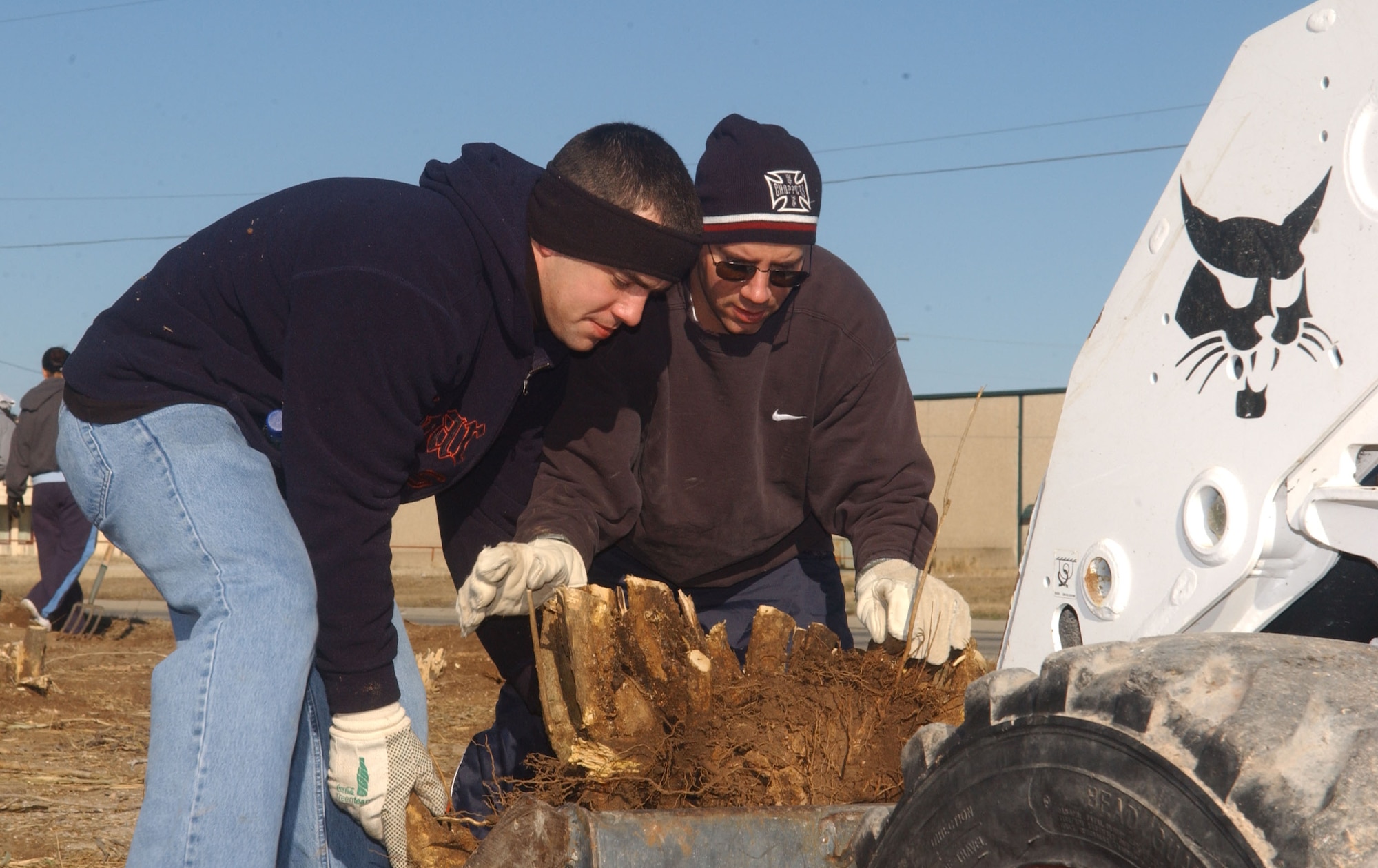 Staff Sgt. Edward Struckle (left) and Tech. Sgt. Brandon O’Neil load the remaining part of a tree trunk onto the lift of a bobcat, a small construction vehicle similar to a bulldozer. (U.S. Air Force photo by Staff Sgt. Gina O’Bryan)