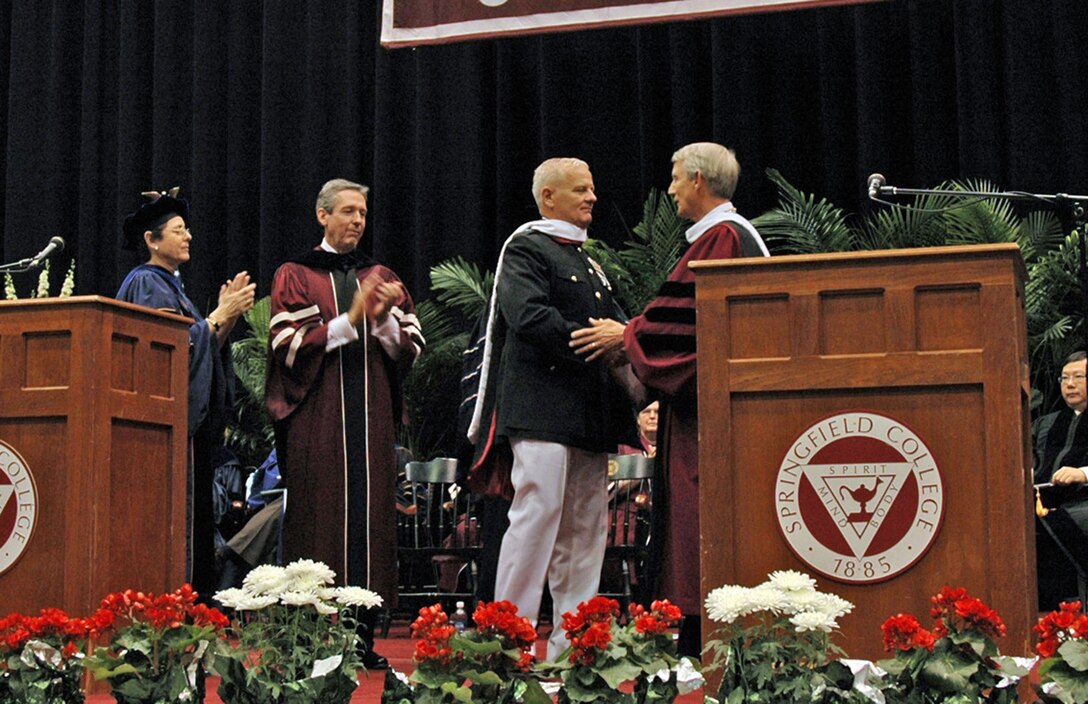 Brig. Gen. Paul E. Lefebvre shakes hands with Dr. Richard Flynn president of Springfield College while Jean A. Wyld vice president for academic affairs and James E. Walsh Chair of Springfield College's board of trustees applaud from left to right respectively. Springfield College is about the spirit of giving and service to others for the college to recognize those values in our Corps values is truly an honor said Lefebvre.