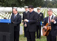 Retired Navy Cmdr. John Alexander, director of communications and public affairs, Navy-Marine Corps Relief Society, reads the roll call of remembrance - the names of the 17 sailors killed in the Oct. 12, 2000, terrorist attack on the USS Cole. Ben Barbin (center) strikes the triangle after each name, as retired Navy Capt. William Perry strums his guitar honoring the dead sailors. Photo by Rudi Williams