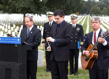 Retired Navy Cmdr. John Alexander, director of communications and public affairs, Navy-Marine Corps Relief Society, reads the roll call of remembrance - the names of the 17 sailors killed in the Oct. 12, 2000, terrorist attack on the USS Cole. Ben Barbin (center) strikes the triangle after each name, as retired Navy Capt. William Perry strums his guitar honoring the dead sailors. Photo by Rudi Williams