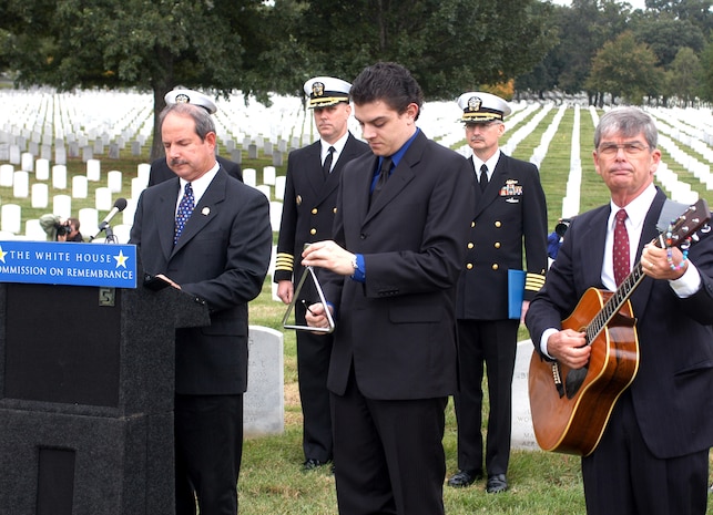 Retired Navy Cmdr. John Alexander, director of communications and public affairs, Navy-Marine Corps Relief Society, reads the roll call of remembrance - the names of the 17 sailors killed in the Oct. 12, 2000, terrorist attack on the USS Cole. Ben Barbin (center) strikes the triangle after each name, as retired Navy Capt. William Perry strums his guitar honoring the dead sailors. Photo by Rudi Williams