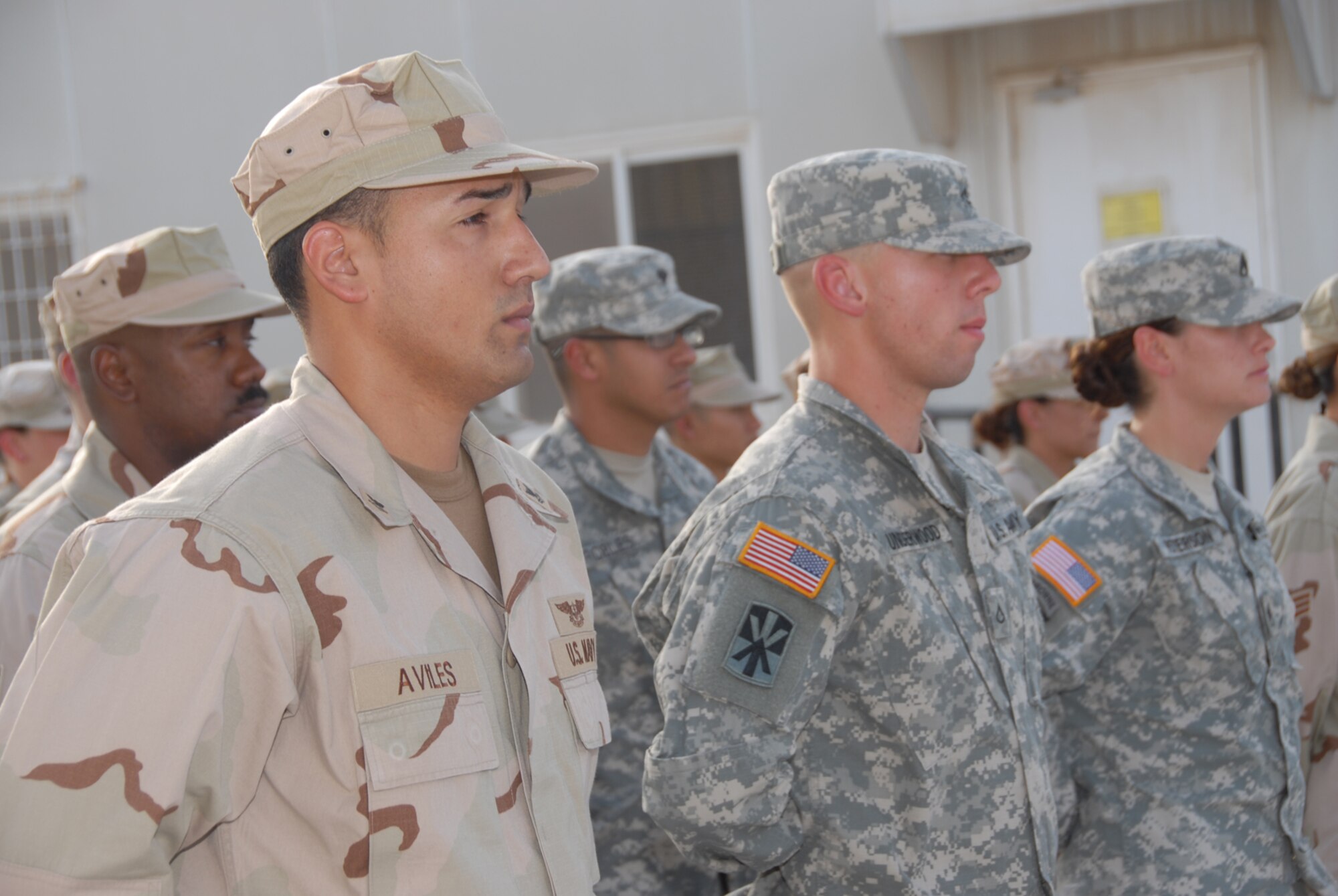 The Desert Eagle Team, comprised of members fo the Air Force, Army, Navy and Marine Corps, stand at parade rest while the 379th Air Expeditionary Wing Honor Guard lowers the flag during reveille at the flag poles near Memorial Plaza in Coalition Compound. The base participated in several activities throughout the day to include; a triathalon, barbecue, darts, limbo and live entertainment. (U.S. Air Force photo by Airman 1st Class Ashley Tyler)