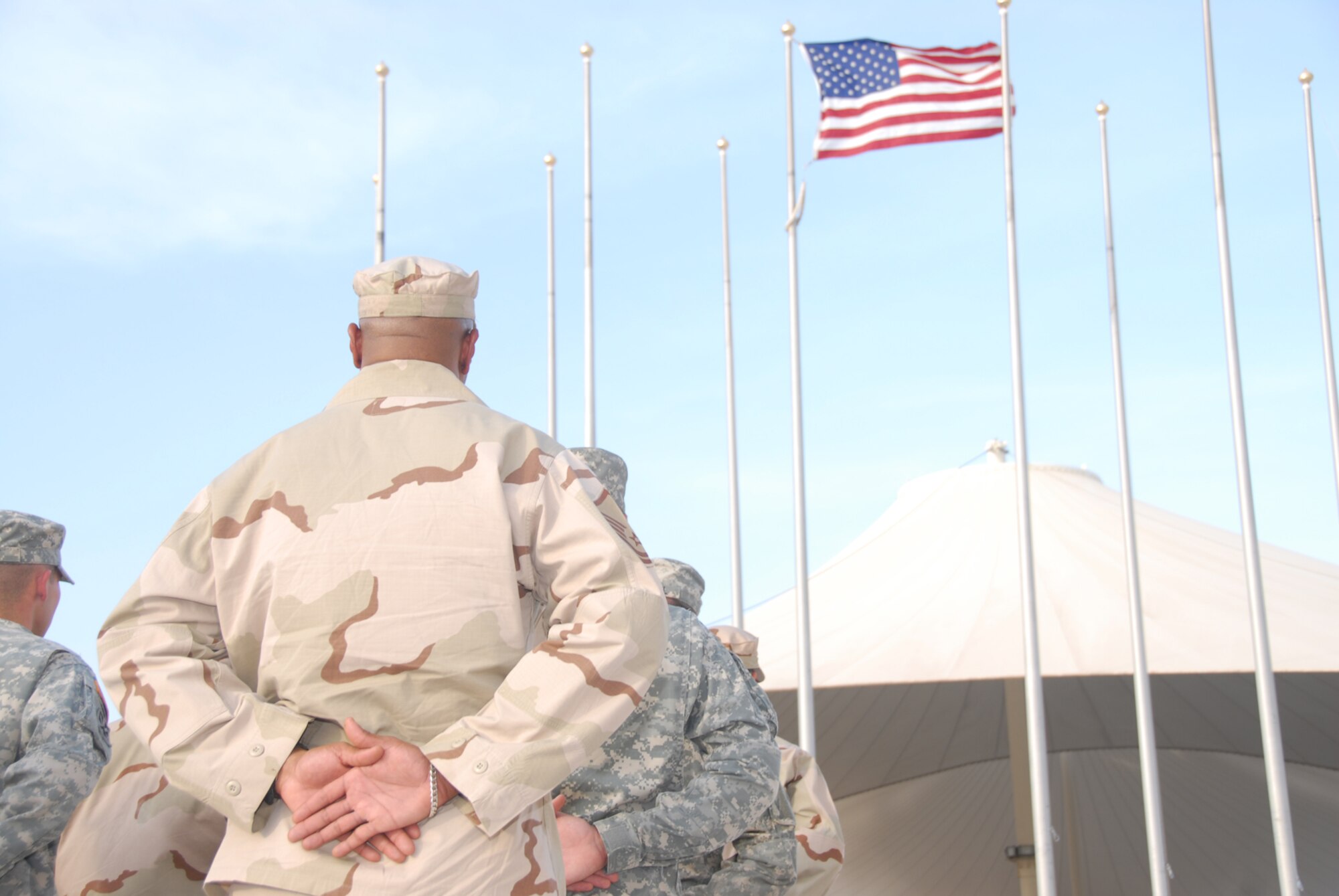The Desert Eagle Team, comprised of members fo the Air Force, Army, Navy and Marine Corps, stand at parade rest while the 379th Air Expeditionary Wing Honor Guard lowers the flag during reveille at the flag poles near Memorial Plaza in Coalition Compound. The base participated in several activities throughout the day to include; a triathalon, barbecue, darts, limbo and live entertainment. (U.S. Air Force photo by Airman 1st Class Ashley Tyler)