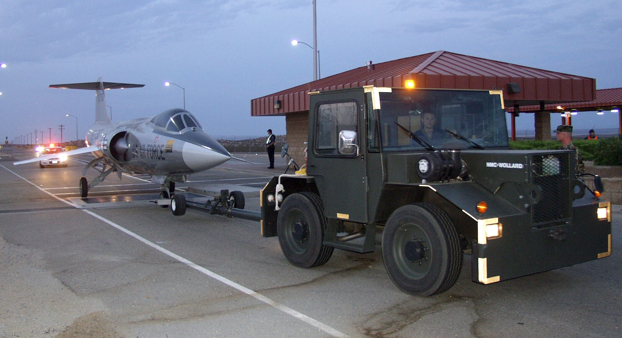 An aircraft tug from the 412th Aircraft Maintenance Squadron tows the last on-base F-104 Starfighter through the outbound lanes of the West Gate on Monday evening. The F-104 was positioned at the Century Circle display outside the gate. The display is scheduled to open in late August. (Photo by Doug Nelson)