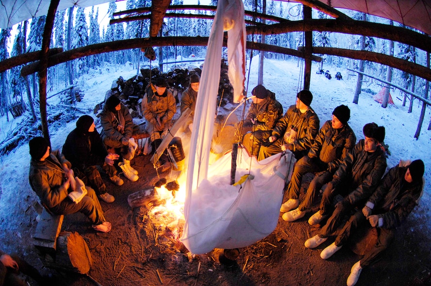 Sitting by a fire to keep warm, students also tend to the water bucket, which is a parachute filled with snow. It hangs near the fire so that the snow will melt and drip into a bucket for drinking water. (Photo by Staff Sgt. Matthew Hannen)