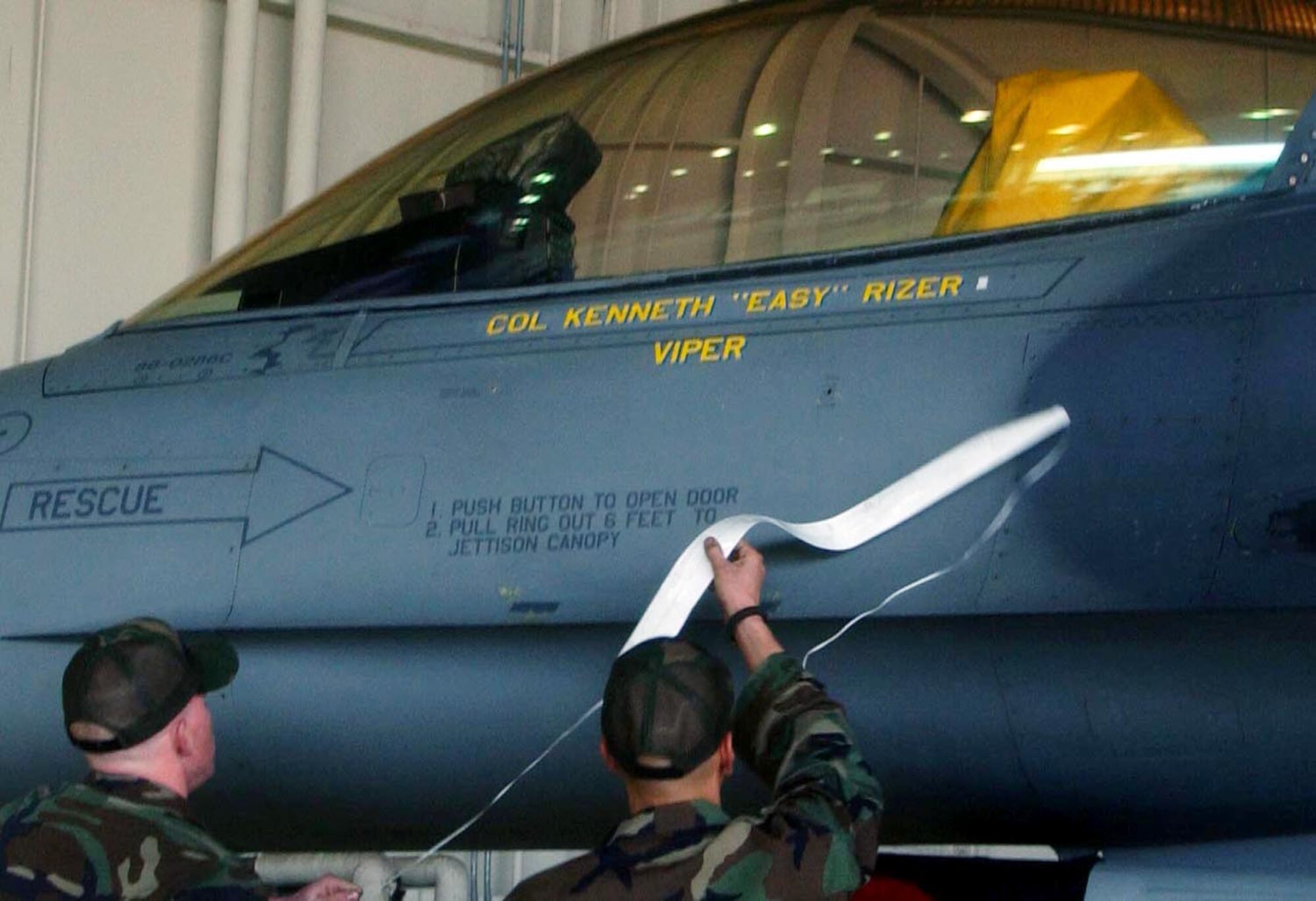 Airman 1st Class Matthew Pavlin and Staff Sgt. Matthew Perry, both F-16 Crew Chiefs from the 80th Aircraft Maintenance Unit, pull a magnetic strip off of the 8th Operations Group flagship aircraft to expose Col. Kenneth "Viper" Rizer's name dedicating it to him while under his command for the next year. Col. Rizer took command of the 8th OG during a ceremony in Hangar 3 here today. (U.S. Air Force photo/Senior Airman Steven Doty)