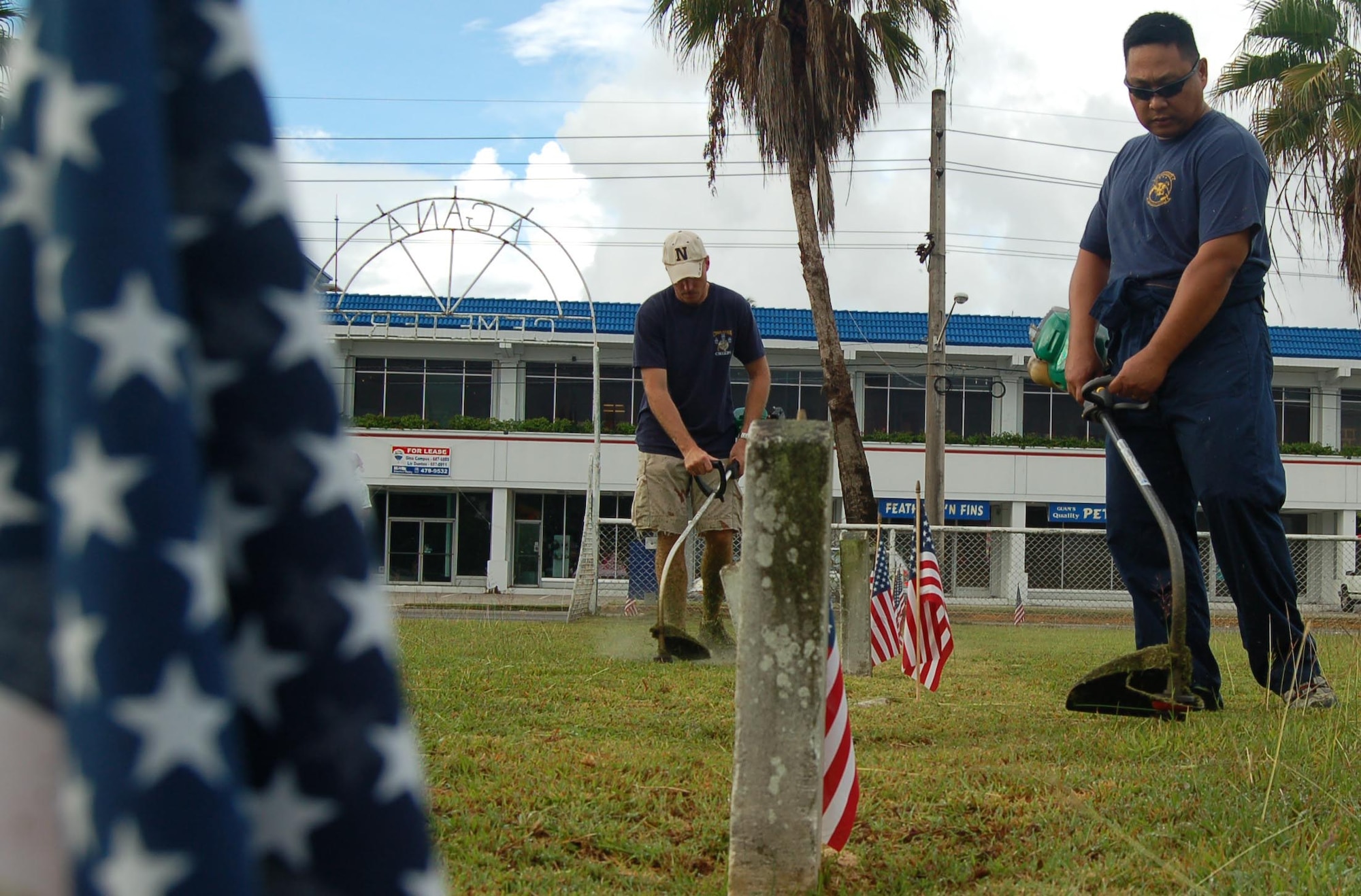Flags adorn the graves of fallen heroes as Navy ATCS Brian O'shea, in the foreground, and Navy SKC Edwin White, both from Helicopter Sea Combat Squadron-Two Five, trim the grass at the Agana Cemetery May 25 as part of the squadron's Memorial Day tribute.  HSC-25 adopted the cemetery in September 2004 after discovering it was a cemetery dedicated to Naval personnel.  Sailors from the HSC-25 care for the cemetery every two weeks.  (Photo by Air Force Public Affairs Tech. Sgt. Brian Bahret/36th Wing Public Affairs)