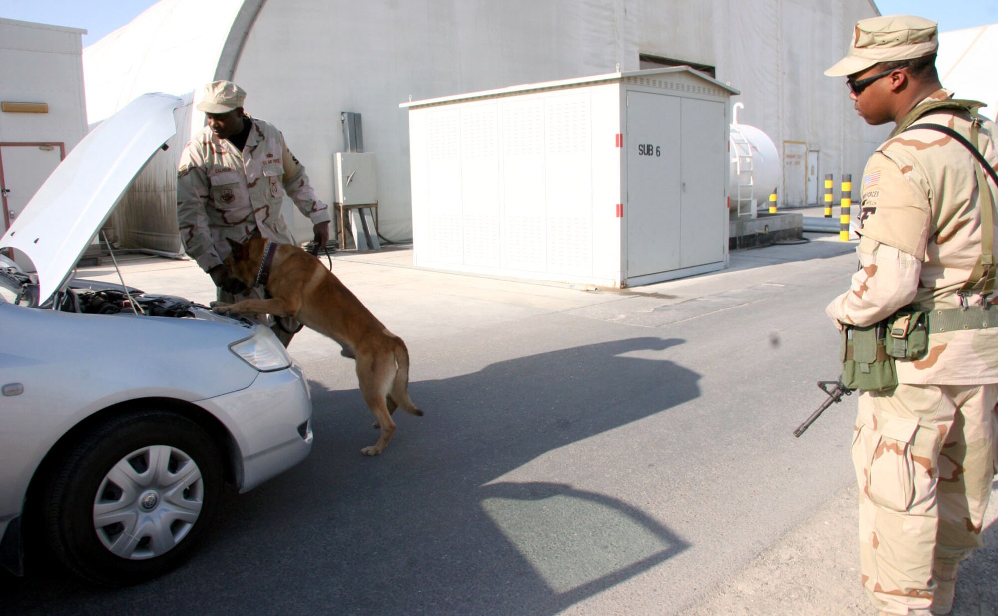 Staff Sgt. Malik Calhoun, 380th Expeditionary Security Forces Squadron K-9 handler, and Arco, his Belgian Malinois patrol and explosive detection dog, search under the hood of a car during a vehicle sweep while his brother Airman 1st Class Dandre Bufford, 380th ESFS provides overwatch during an explosive detection training scenario Nov. 27. (U.S. Air Force photo/Master Sgt. Ruby Zarzyczny) 