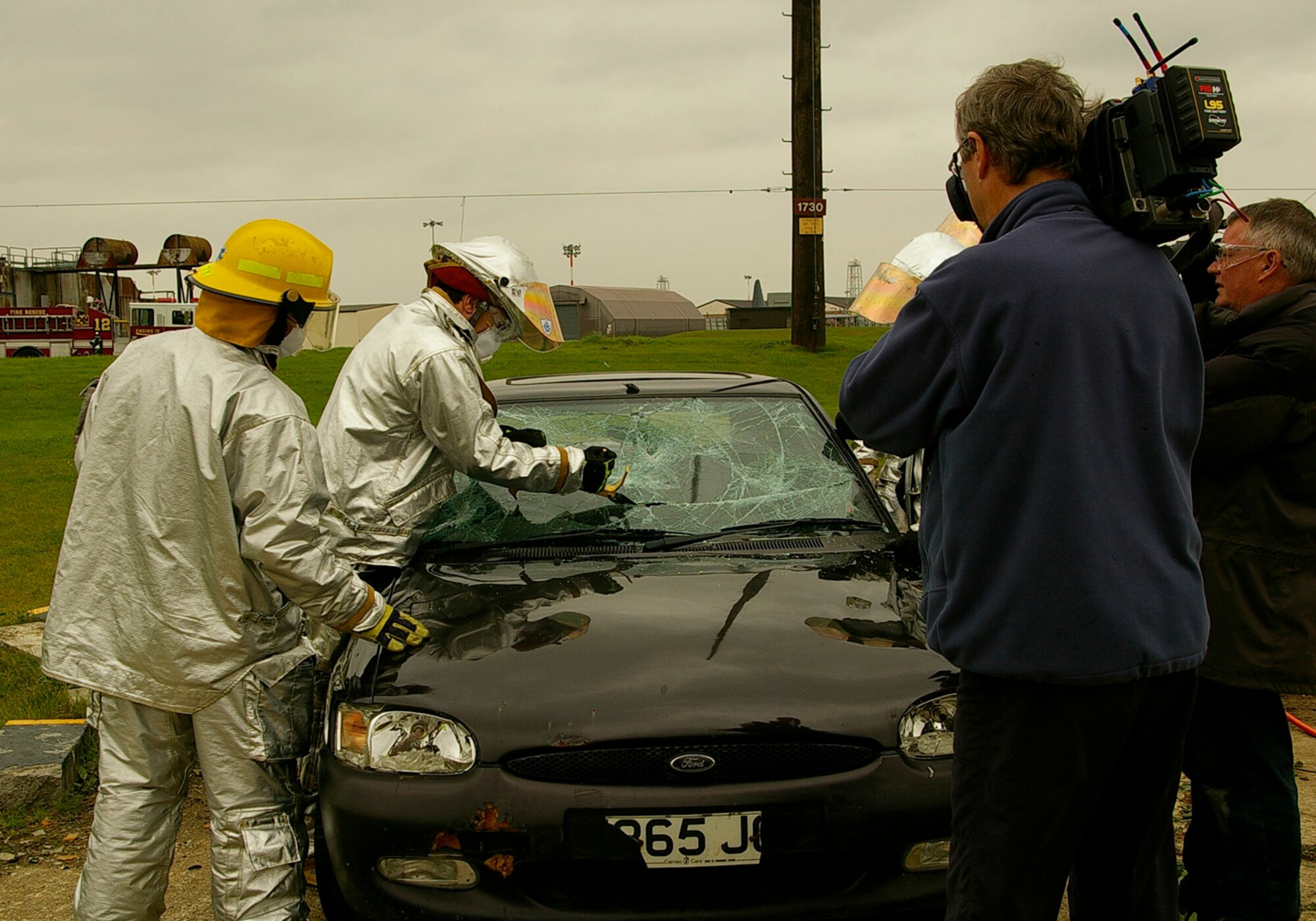 Tech. Sgt. Richard Henderson, left, 100th Civil Engineer Squadron Fire Department, keeps a close eye on Gethin Jones, center, presenter of children's TV show, "Blue Peter," as he helps cut the windshield out of a car. Gethin was at RAF Mildenhall Oct. 1 and 2 and was being filmed training with the firefighters for an upcoming edition of the show. (U.S. Air Force photo by Karen Abeyasekere)