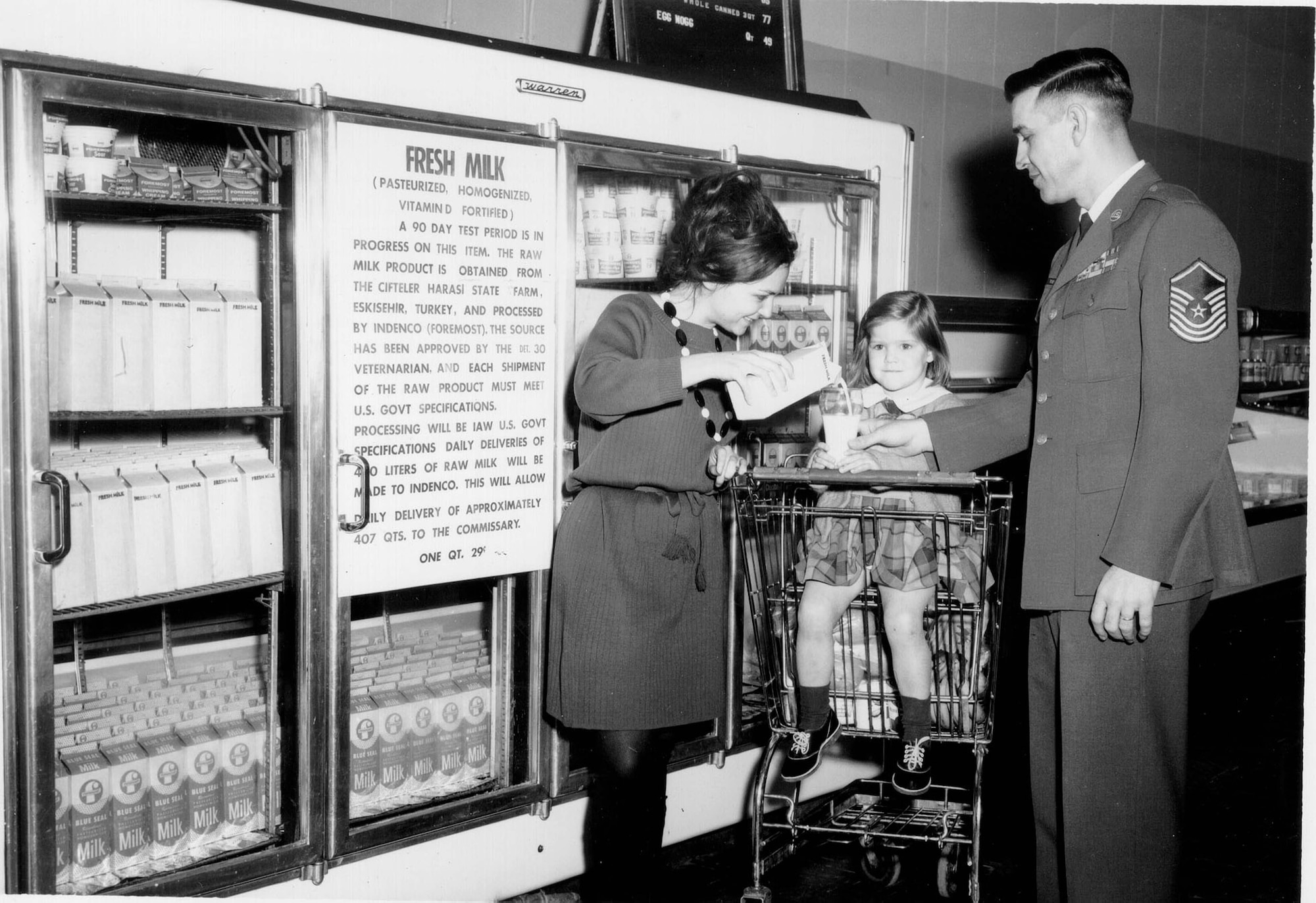 In this 1967 photo, parents pour a glass of fresh milk for their daughter inside the Ankara Air Base, Turkey, commissary. This was a publicity photo for a 90-day test program for daily deliveries of 407 quarts of fresh milk. The milk was obtained from a Turkish state farm meeting U.S. standards, and was processed by a contractor following American specifications. The poster on the cooler says the finished product was “Pasteurized, Homogenized [and] Vitamin D fortified.” The program exemplifies the special concerns and needs of families living overseas at the far end of a long supply pipeline. (DeCA historical file) 
